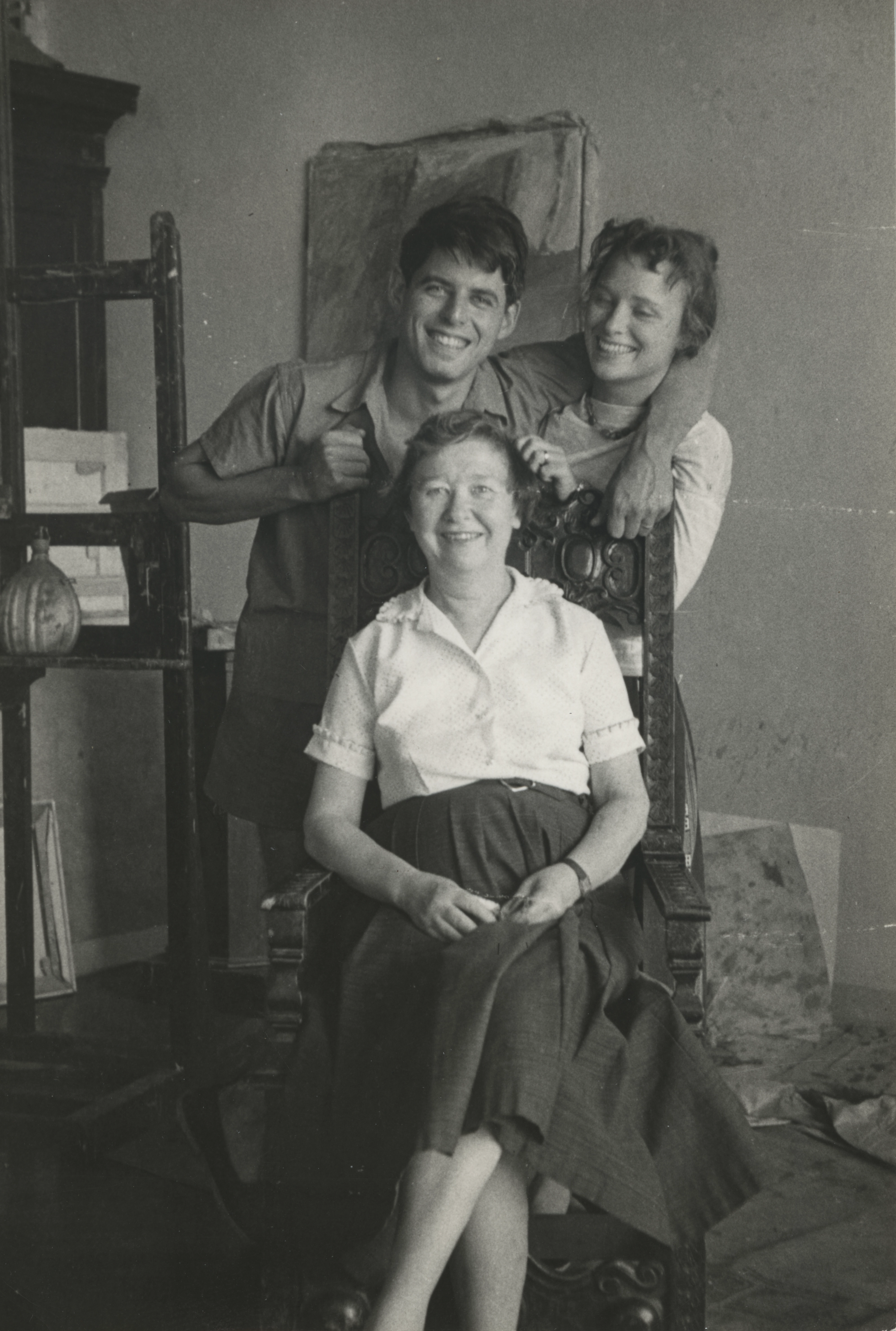 Black and white photograph from 1958 of three people with light skin tones; a younger man and younger woman, standing behind a chair, and an older woman seated in front of them. All three are smiling.