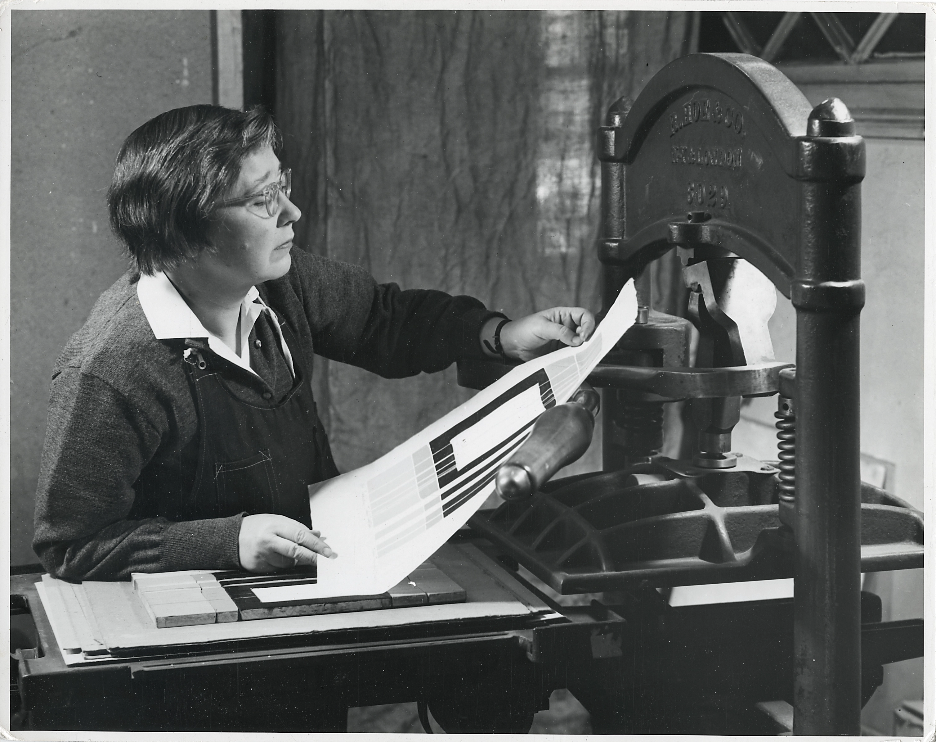 Black and white photograph from 1954 of a light-skinned, middle aged women, examining a print just pulled off of a large printmaking press.