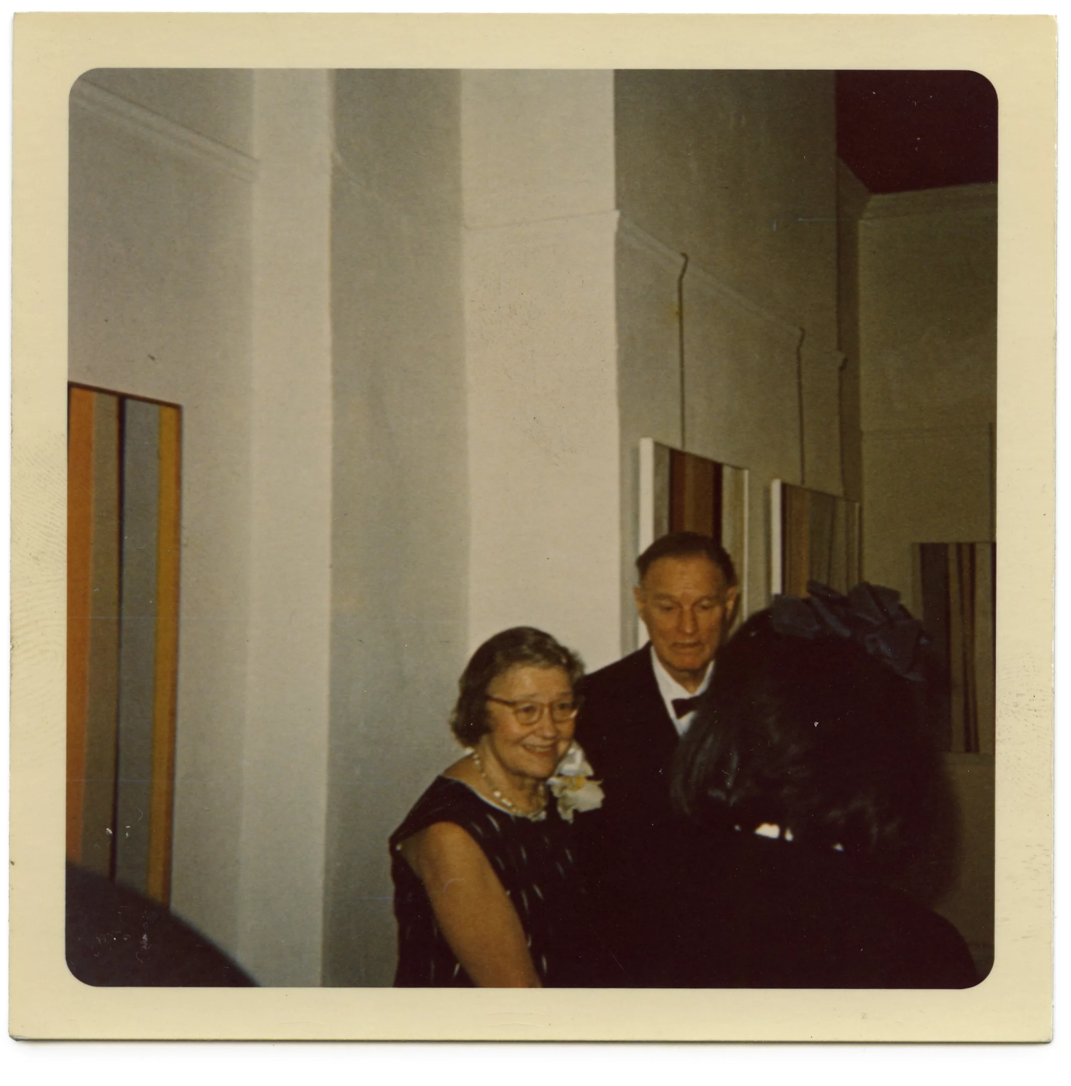 Photograph of a man and a woman, both with light skin tones, standing in front of abstract paintings.