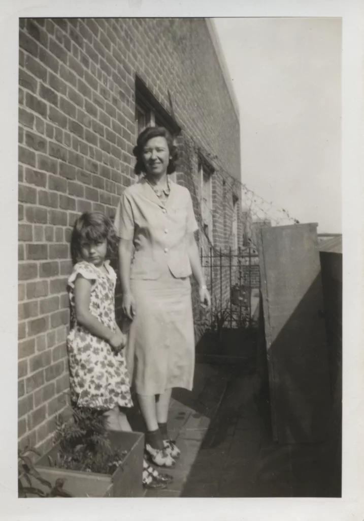 Black and white photograph of a young girl and a young woman, both with light skin tones, standing on an NYC rooftop against a brick exterior wall.