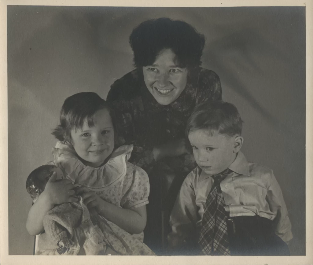 Black and white portrait from the late 1930s of two seated children and a women standing over them, all with light skin tones.
