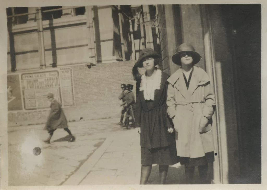Black and white photograph from 1922 of two young women, both with light skin tones, dressed in 1920's fashion. They stand in an outdoor plaza, with three police officers seen in the background.