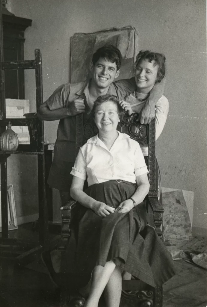 Black and white photograph from 1958 of three people with light skin tones; a younger man and younger woman, standing behind a chair, and an older woman seated in front of them. All three are smiling.