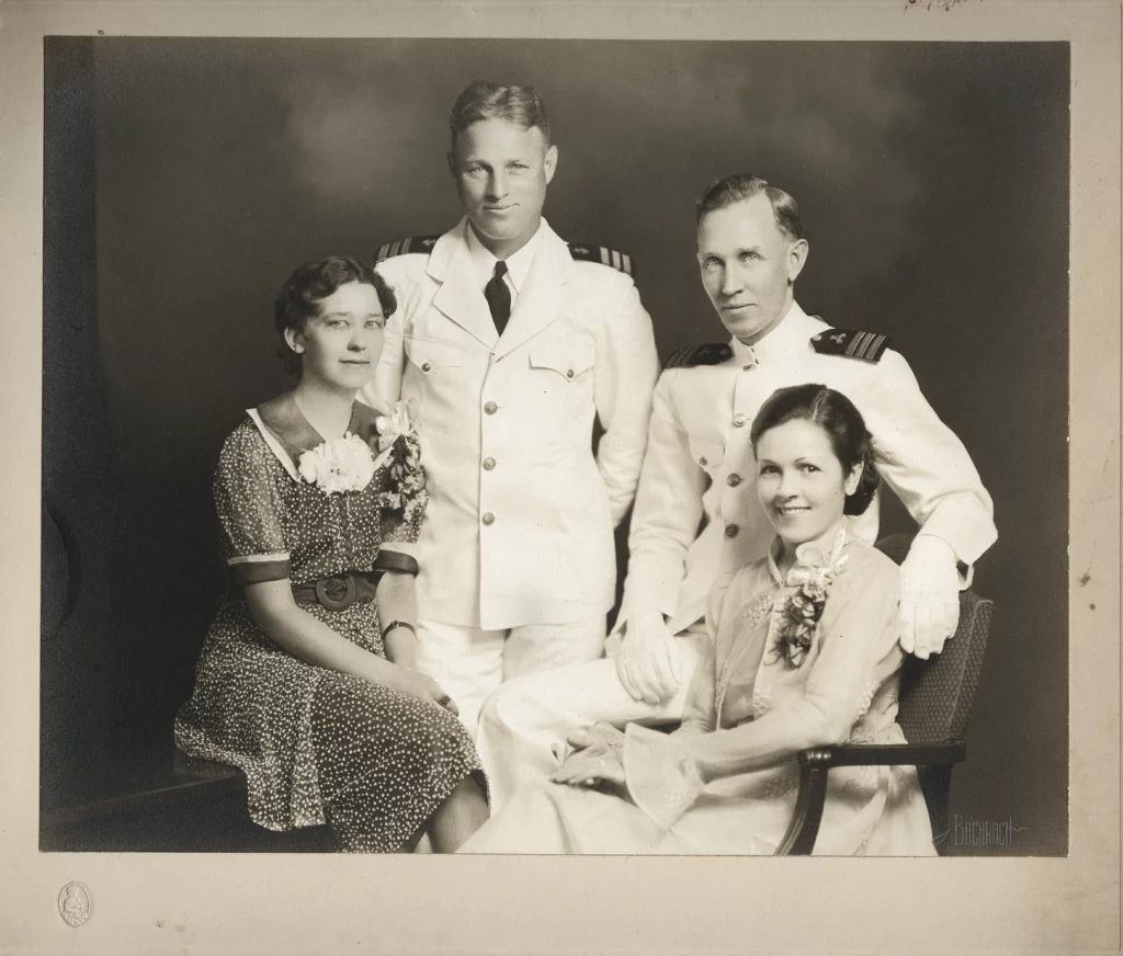 Black and white photograph from 1929 of two men in white navy uniforms and two seated women formally dressed, all with light skin tones.