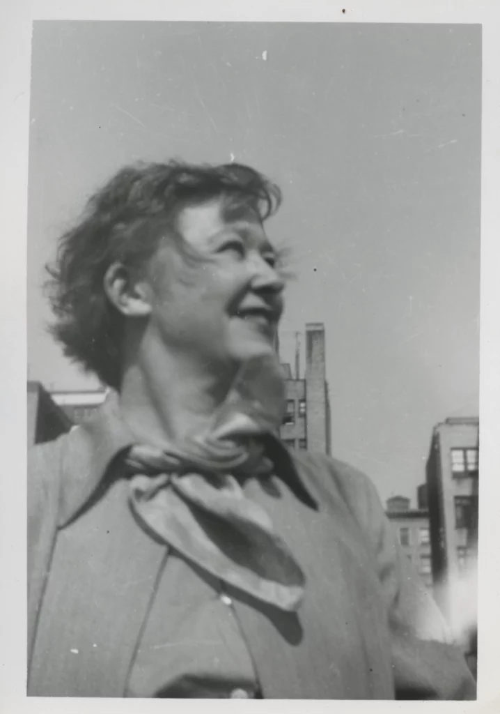Black and white portrait of a woman with a light skin tone, looking off to the right and smiling; NYC apartments are seen behind her.