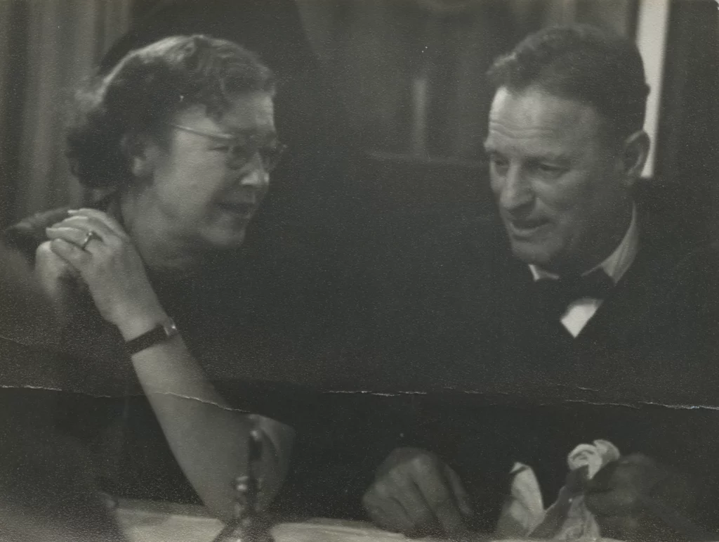 A black and white photograph from the last 1950s of a woman and man, both with light skin tones, formally dressed and seated a table in conversation.