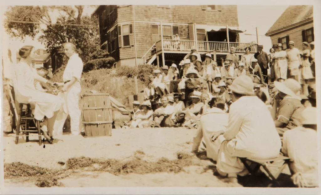 Sepia tone photograph from 1927 of a group of students seated outdoors, observing two people next to a canvas.