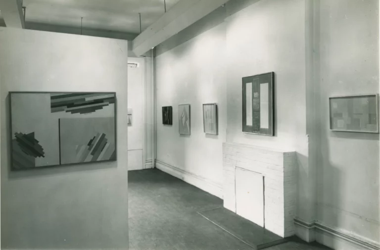 Black and white photograph of the interior of a white-walled gallery space, with abstract paintings hung on the walls.