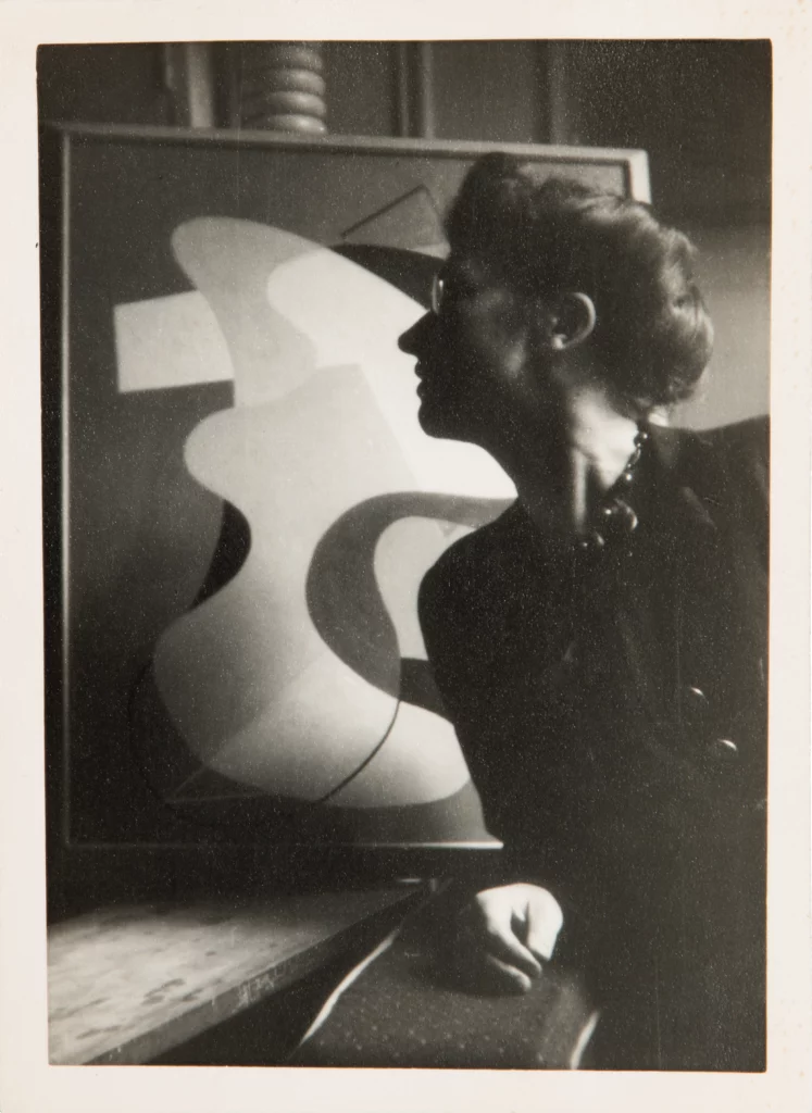 A 1942 black and white side portrait of a woman with a light skin tone and glasses, in front of an abstract painting.