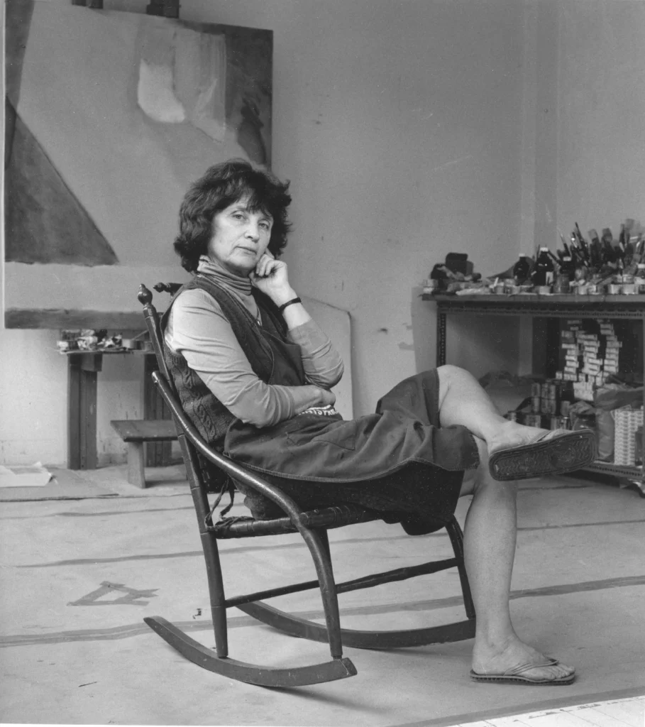 Black and white photograph of a light-skinned woman with short hair, sitting a rocking chair in a painting studio.