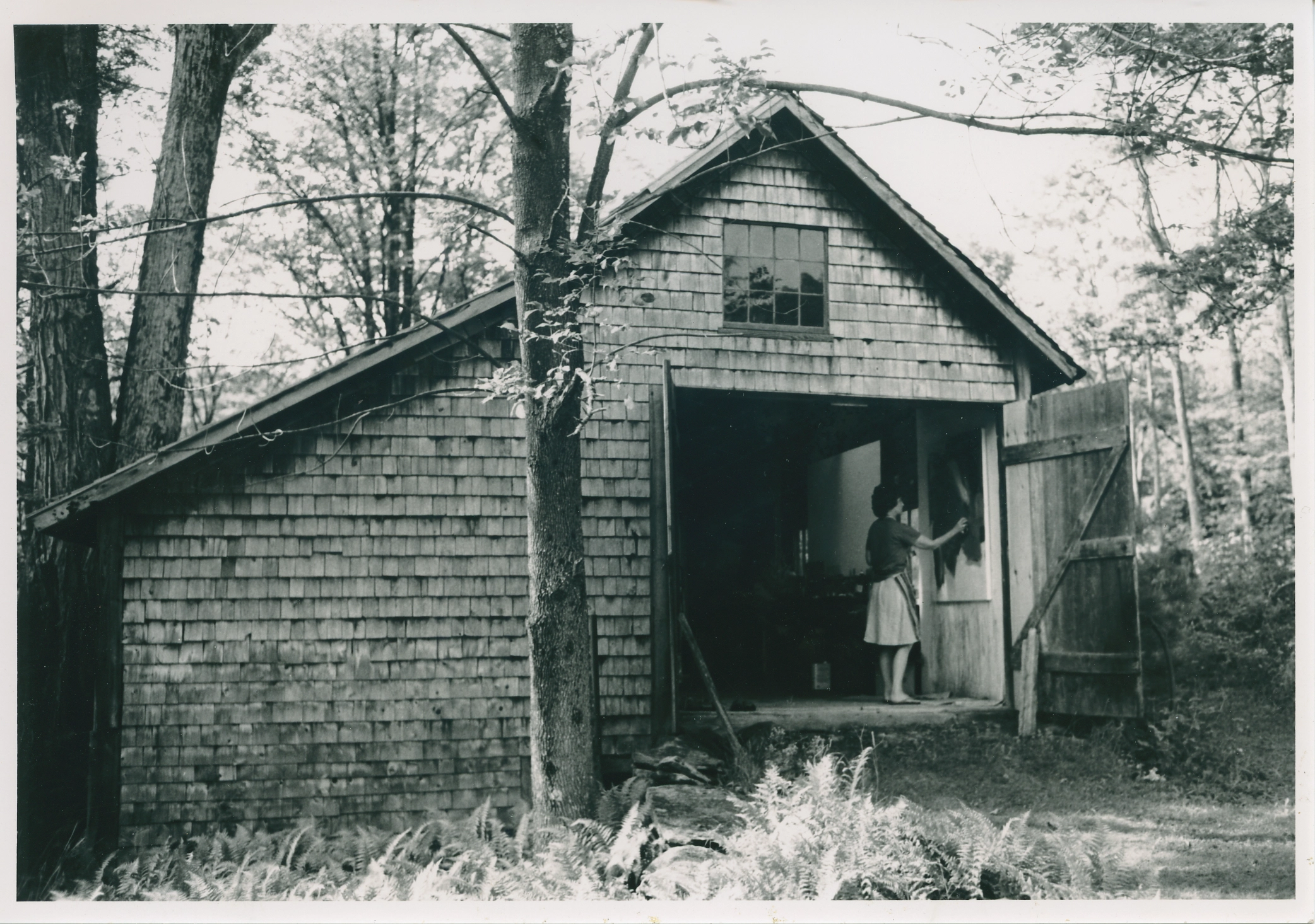 Black and white photograph of barn in the woods among trees and plants. The double barn doors are open, revealing a woman inside, painting on a canvas.