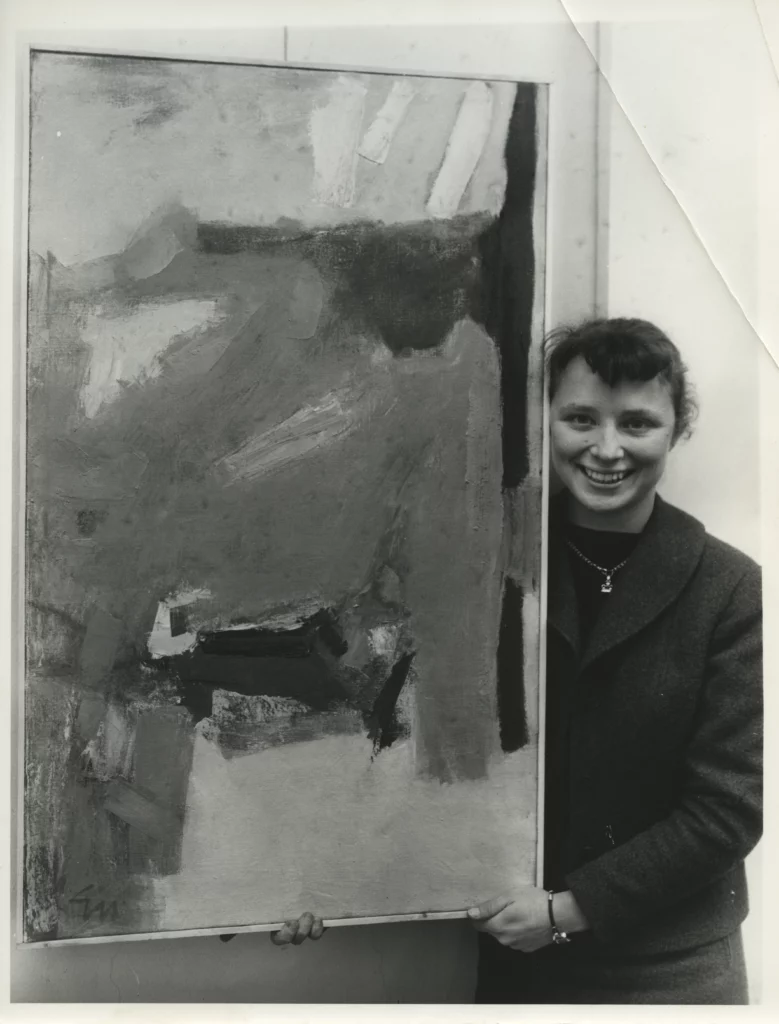 A black and white photograph from the 1960s of a smiling young woman with a light skin tone standing next to, and holding up, an abstract painting.