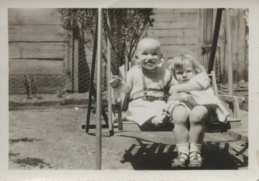 A black and white photograph from the early 1930s of two chillden with light skin tones sitting on a swing outdoors.