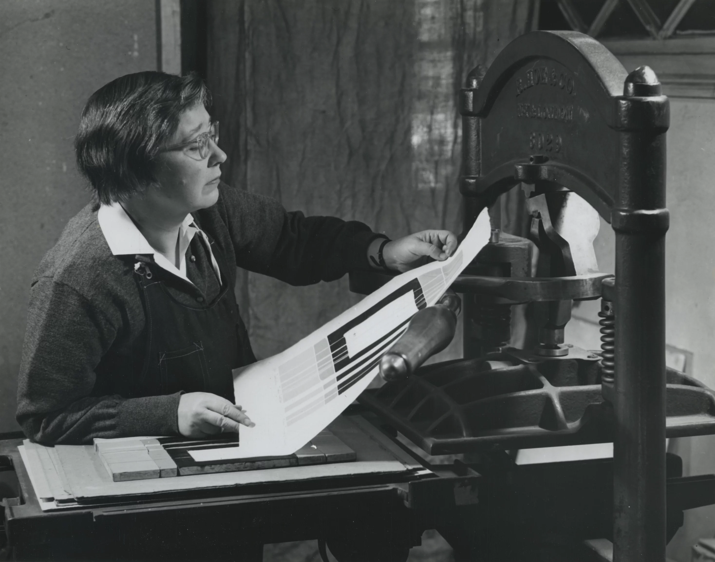 Black and white photograph from 1954 of a light-skinned, middle aged women, examining a print just pulled off of a large printmaking press.