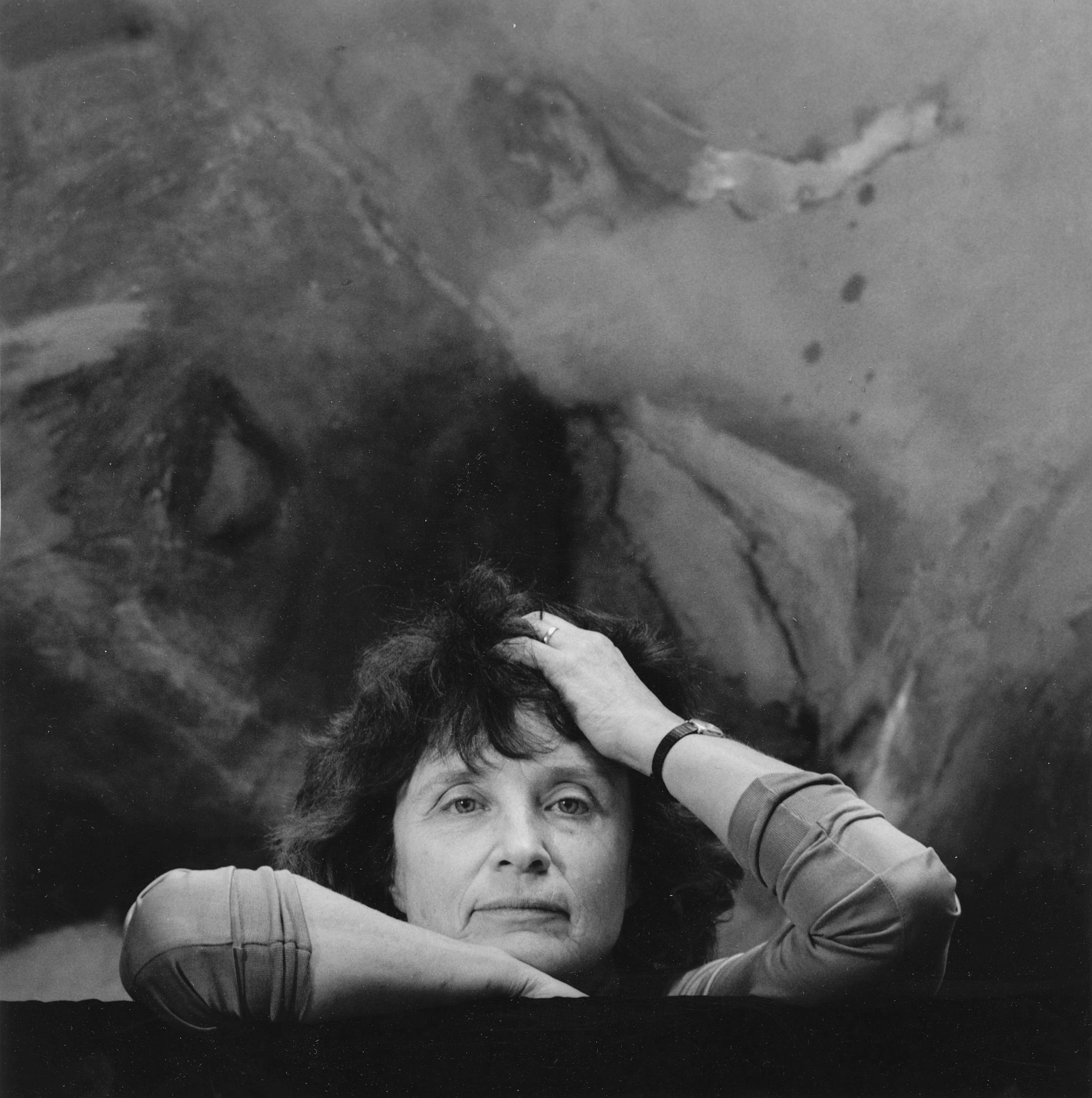 Black and white portrait photograph of a woman with pale skin in front of an abstract painting; just her face and arms are shown as she leans her elbows on a flat surface.