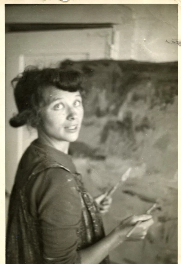 Black and white photograph from 1959 of a woman with a light skin tone, staring back over her right shoulder to the photographer to pause her work on an abstract painting, holding a brush in one hand and a palette knife in another.