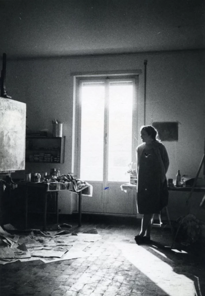 Black and white photograph from 1963 of a woman with a light skin tone standing near a window and looking over to a table of painting supplies and a canvas on a easel.