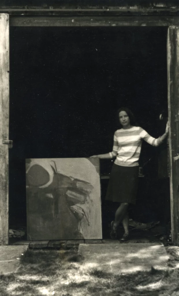 Black and white photograph from 1965 of a woman with a light skin tone, wearing a horizontal striped shirt and long skirt, standing in a barn doorway next to an abstract painting.