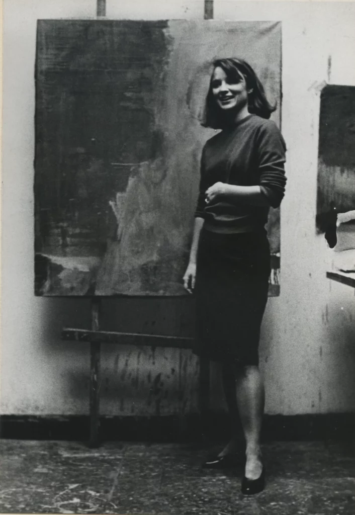 Black and white photograph from 1966 of a young woman with a light skin tone and shoulder-length straight hair standing infant of an abstract painting.