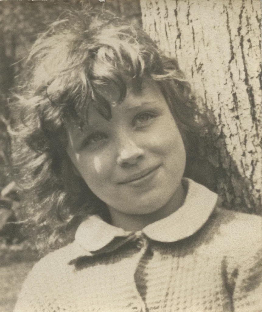 Sepia toned portrait photograph from the mid-1930s of a light skinned girl leaning against a tree outdoors.