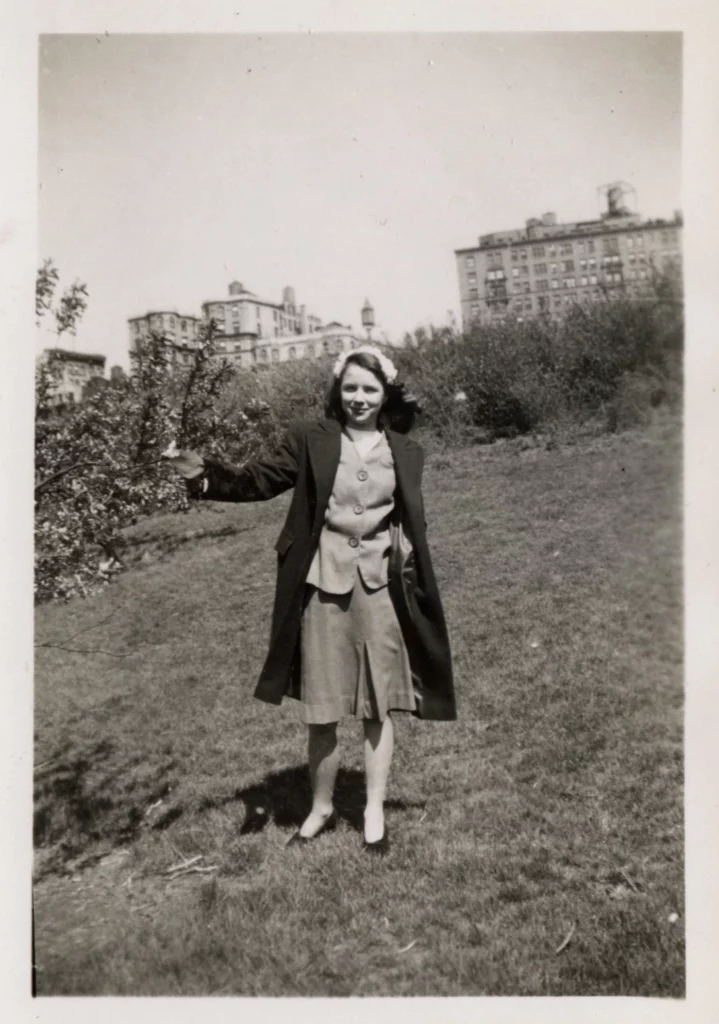 Black and white photograph from the mid-1940s of a young girl outdoors, standing on grass; New York City buildings can be seen in the distance behind her.
