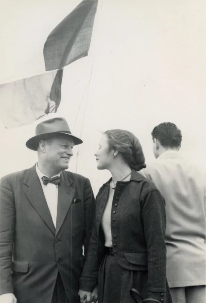 A black and white photograph from the 1950s of an older man and a younger woman, both with light skin tones, looking at each other on a ship. On the right, there is a man with his back turned, looking out onto the water.