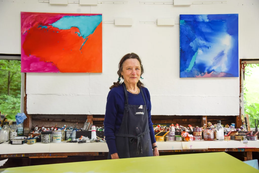 Photograph from 2018 of an older women with a light-skin tone in an art studio, infant of tubes of paint and brushes. Two abstract paintings hang on the wall behind her, a predominantly red on the left, and a predominantly blue on the right.