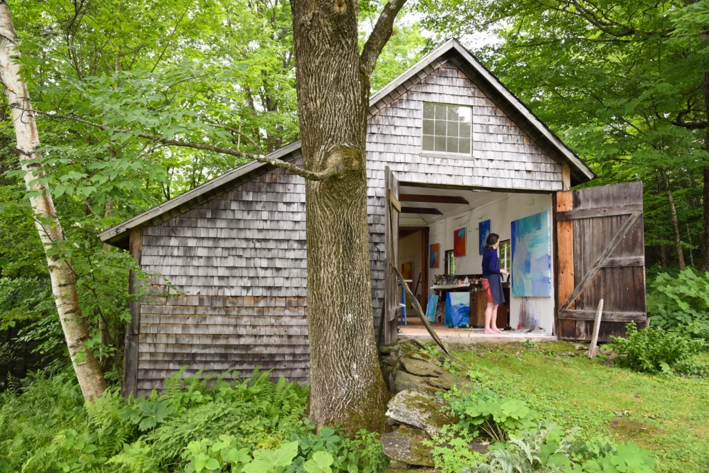 Photograph from 2018 of a brown-shingled barn among the woods and greenery; the doors to the bar are open, revealing an older woman with a light skin-tone working on a light blue abstract painting.