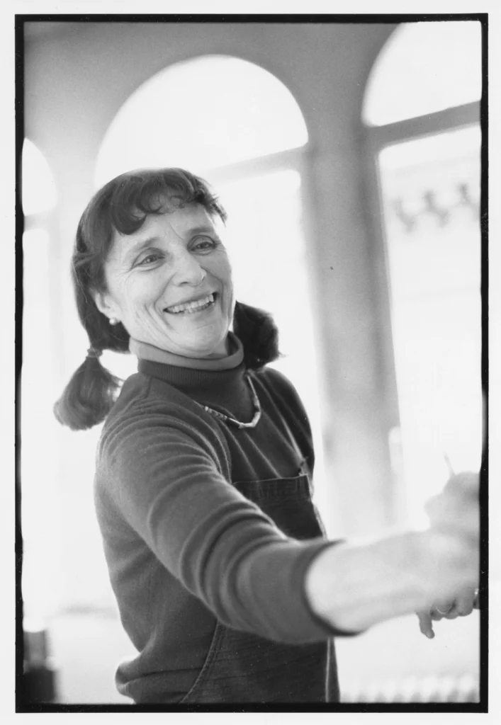 Black and white photograph of a smiling, middle-aged woman with a light skin tone, her right arm outstretched in the process of painting.
