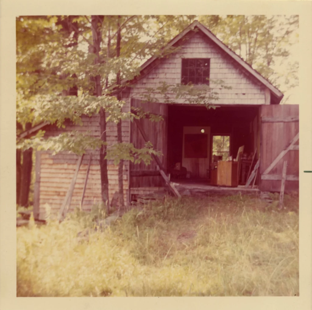 Photography circa 1970s-1980s of a barn with the front doors open, revealing an artist's studio inside. The barn is surrounded by trees and vegetation.