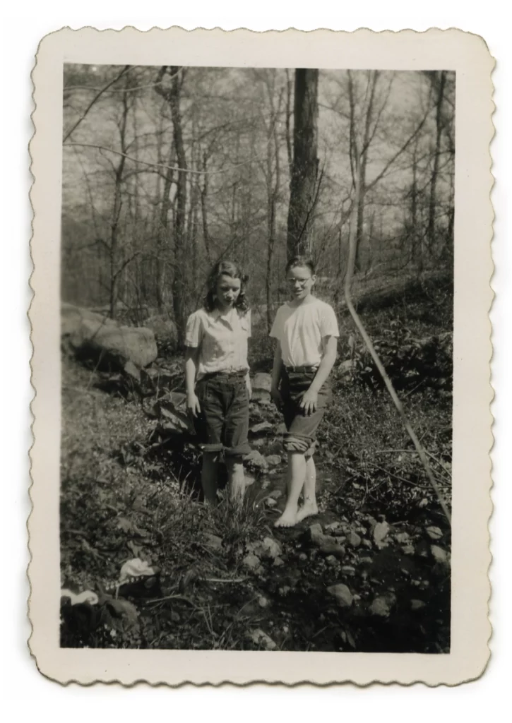 Photograph from the mid-1940s of two middle-school aged children with light skin tones, standing in a creek in the woods. There is a girl on the left, and a boy with glasses on right.