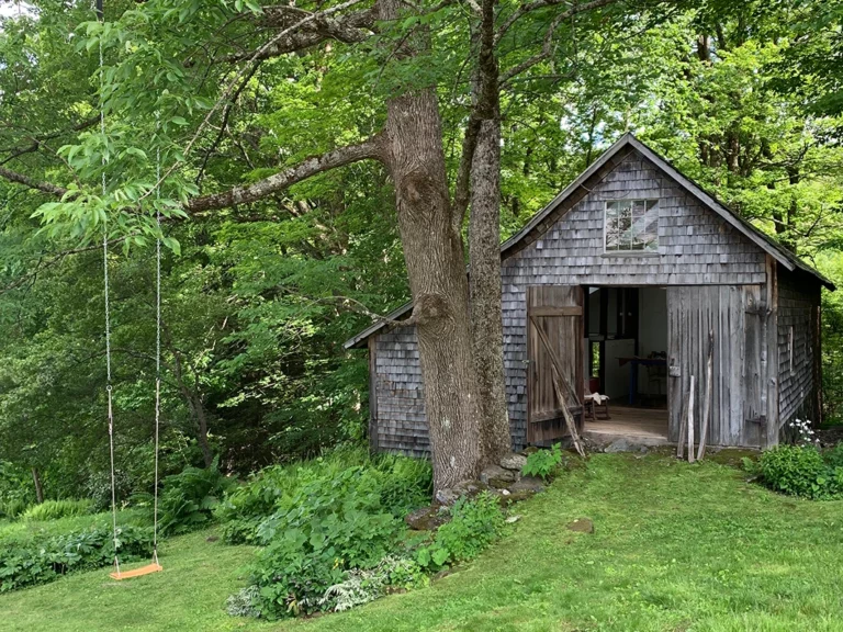 A photograph of a brown barn nestled with it's doors open nestles in lush greenery and woods. A swing hangs from a tree on the left.