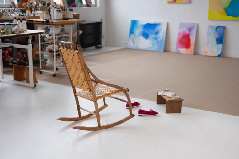 Image of a brown rocking chair on a white floor with a pair of pink slippers and a wooden surface beside it. The far wall from the chair has abstract paintings propped up against it.
