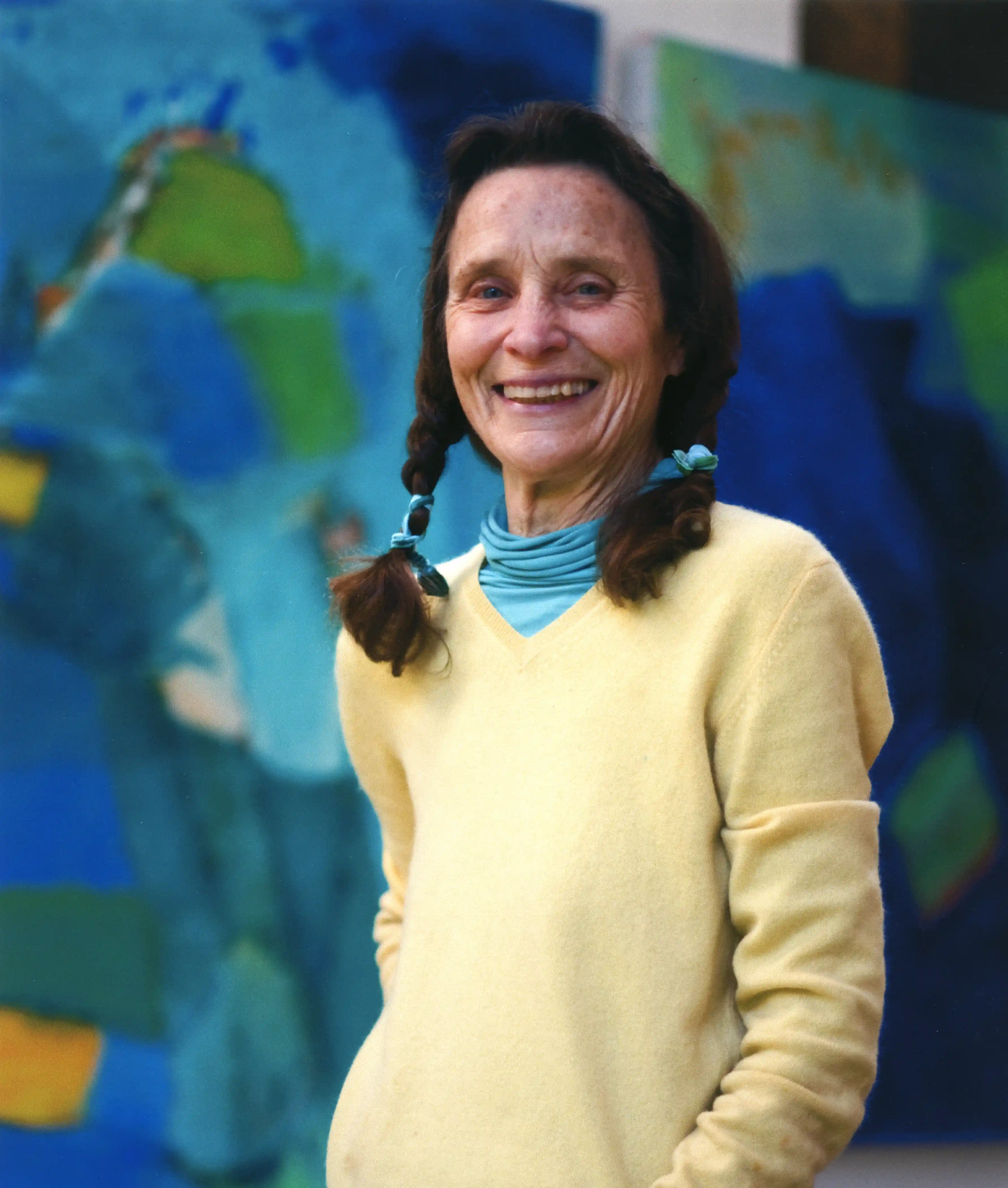 Portrait of a 67 year-old woman with a light skin tone wearing a yellow sweater over a turquoise turtleneck. She is standing infront of two abstract paintings composed of green and blue gestures.