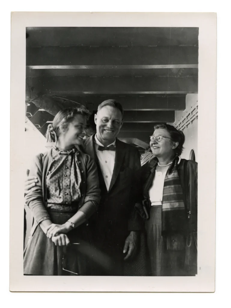 Black and white photograph of a man, young woman, and an older woman on a ship, all with light skin tones and smiling. The two women are looking at each other.