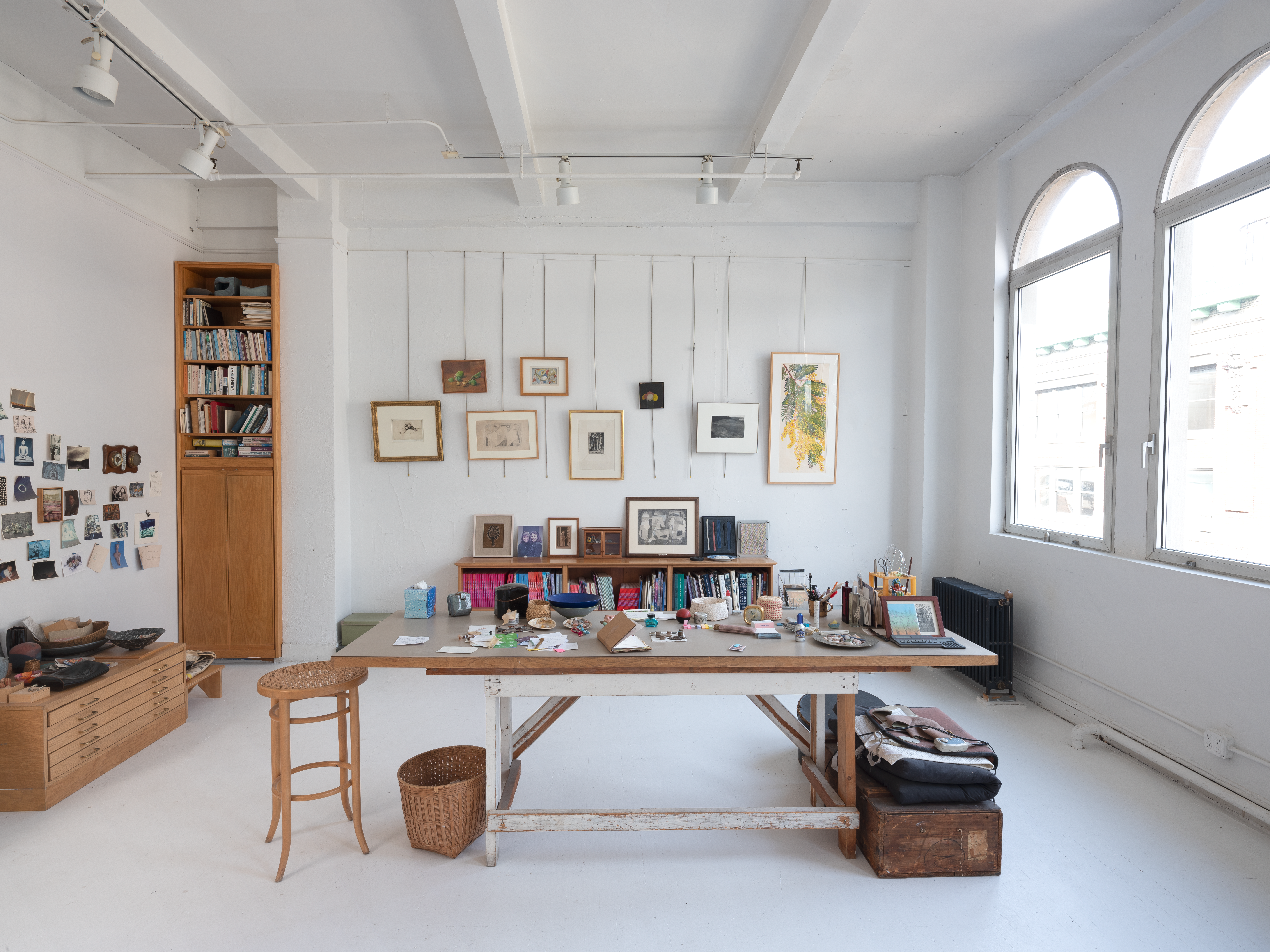 A photograph of a studio room with a table in front of two bookshelves and hanging pictures. To the right, there are two large windows.