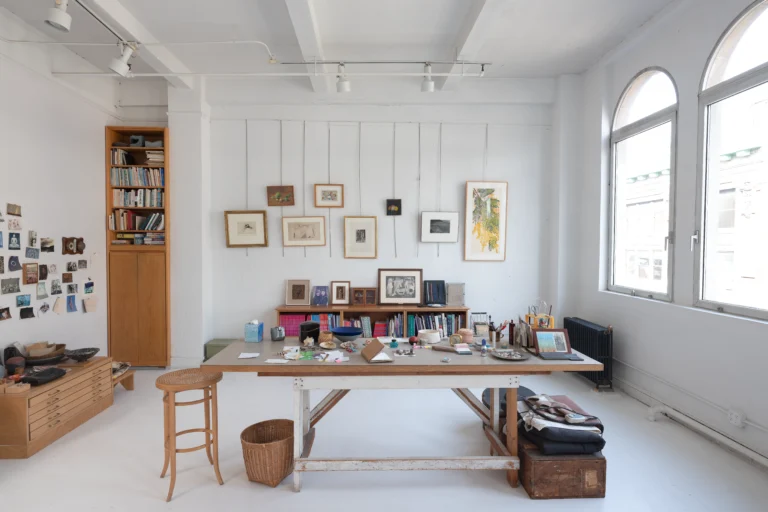 A photograph of a studio room with a table in front of two bookshelves and hanging pictures. To the right, there are two large windows.