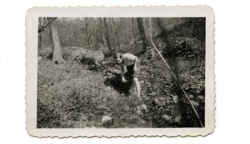 A black-and-white photograph of a young, light-skinned girl in a brook, surrounded by woods.