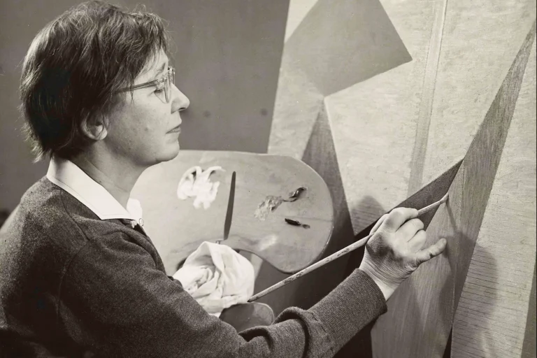 A black and white photograph of a light-skinned woman, holding a palette in her left hand and a paint brush on the right; she is in the process of creating an abstract painting.