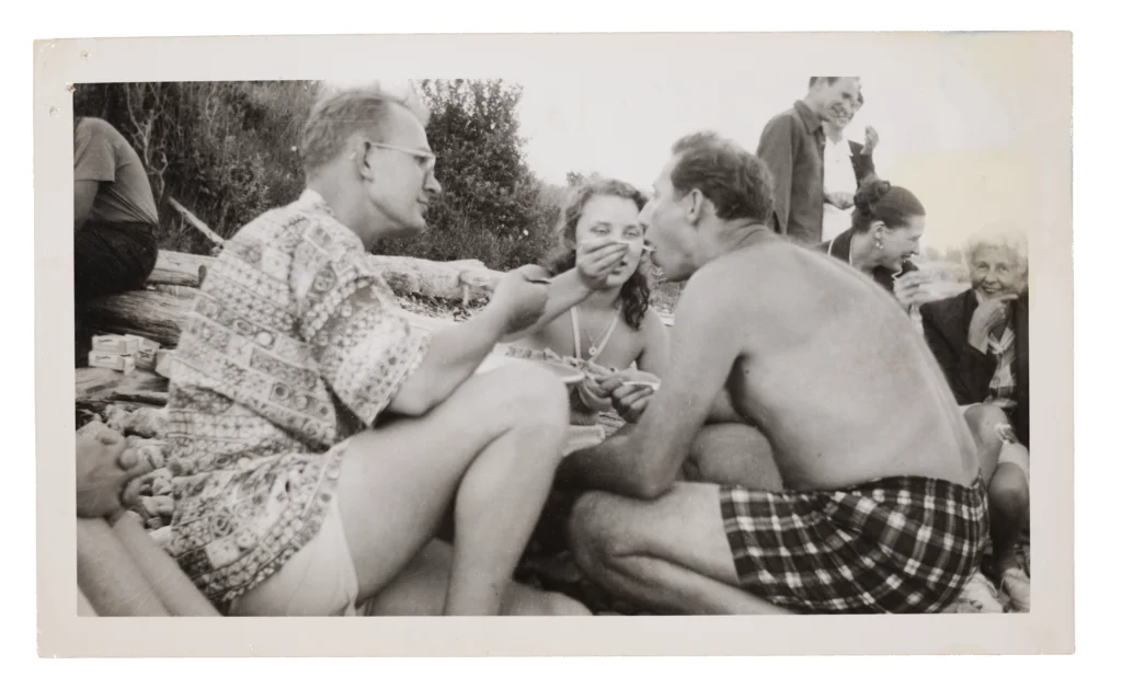 Black and white photograph from the 1950s of a picnic, with two light-skinned men in the foreground — a young adult, light-skinned woman is towards the background, feeding the man on the right with a spoon.
