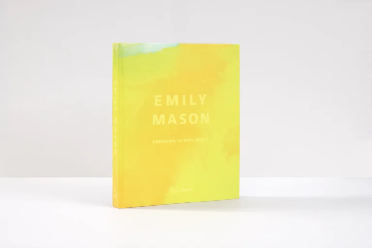 A yellow book cover with the text "Emily Mason: Unknown to Possibility" on the front, in light yellow text. The book is sitting upright on a white table with a white background, and is casting a shadow.
