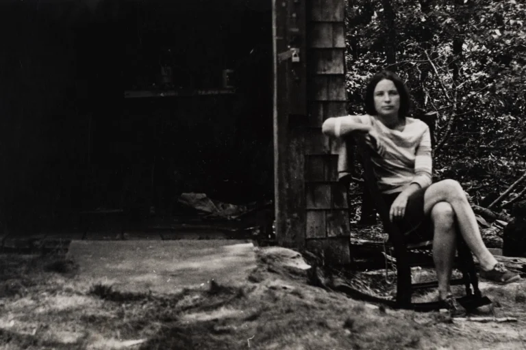 A black and white photograph of a woman sitting sideways in a chair outside of an open barn door.