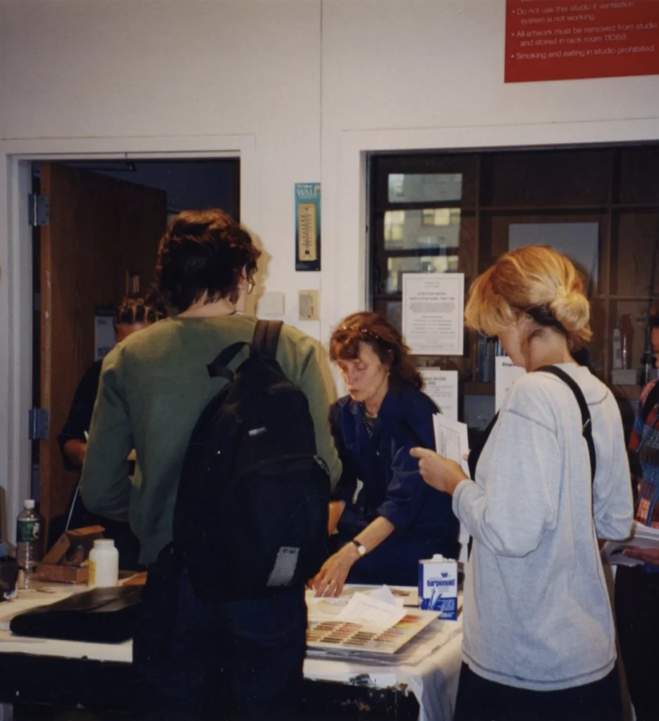 An image of a light-skinned woman with brown hair at a classroom desk with two students in front of her. 