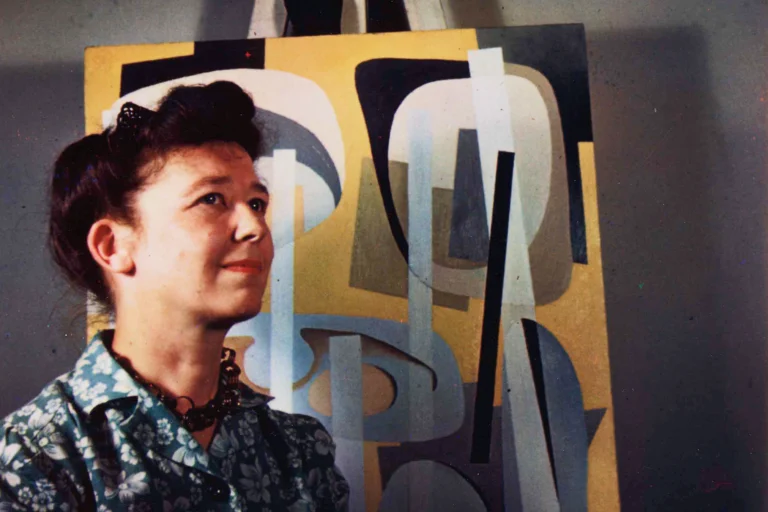 Color portrait photograph from 1946 of a woman with a light skin tone, standing in front of a yellow abstract painting resting on an easel.