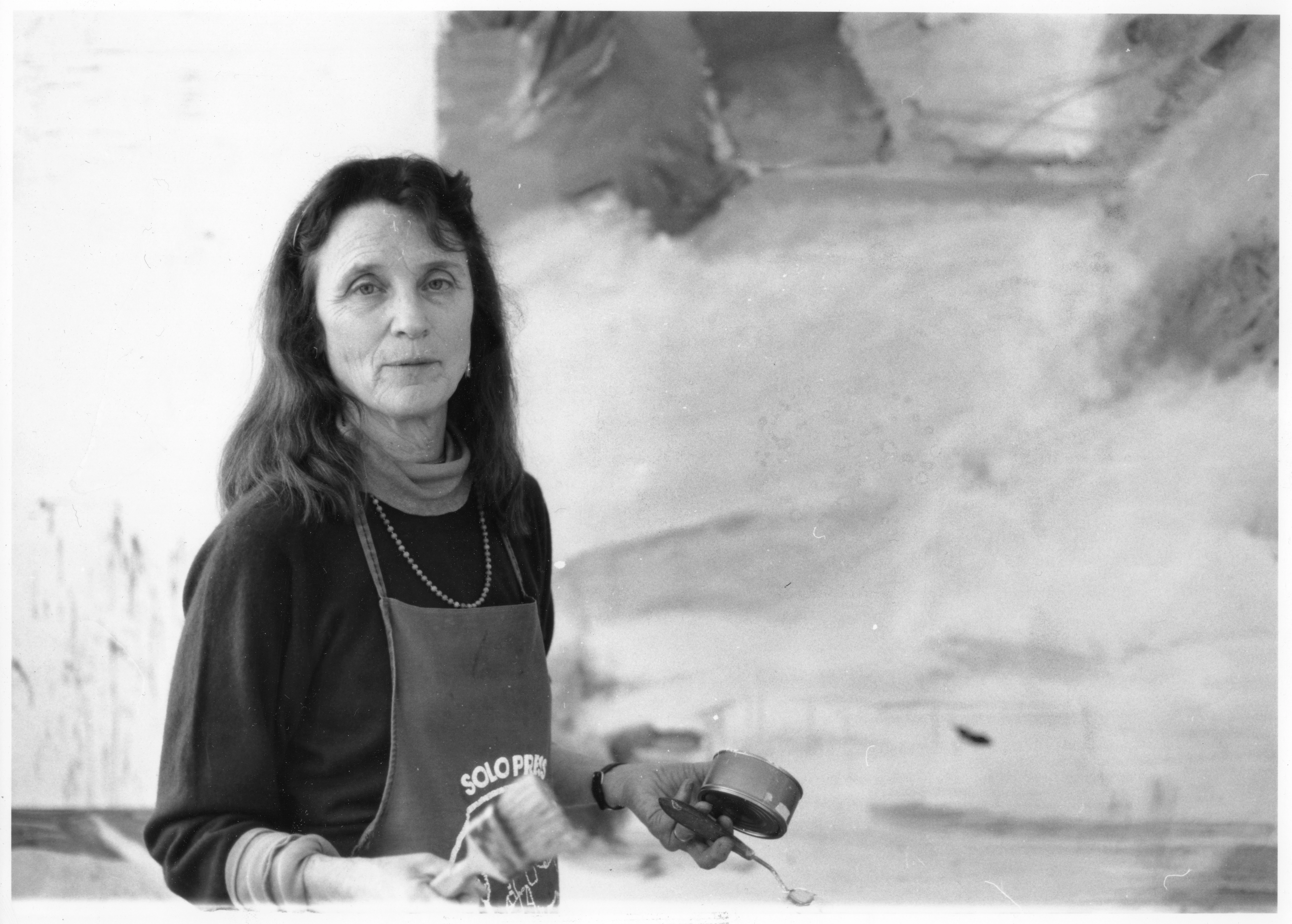 1987c_EM_in_NY_STUDIO_Phong_Bui-01 A black and white photograph of a light-skinned woman looking at the camera., and standing infront of an abstract painting, holding paint cans and brushes.