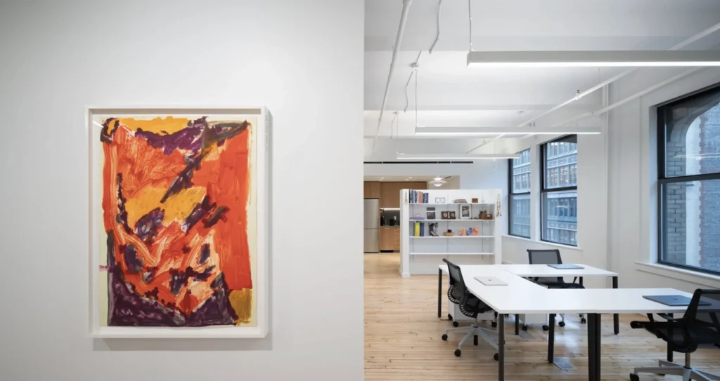 An image of the interior of an office space with light wood floors and white walls. To the left, windows line the wall, and there are office chairs and tables. On the gright, there is an orange and yellow abstract artwork shown hanging on the wall.