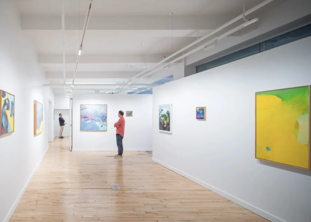An image of the interior of an office space with light wood floors, white walls, and colorful abstract paintings on the walls. Two men are seen in the distance observing the artworks.