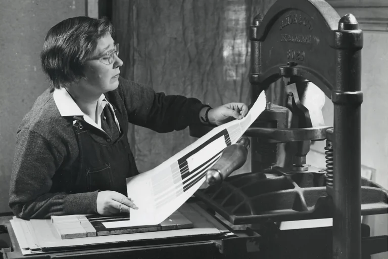 Black and white photograph from 1954 of a light-skinned, middle aged women, examining a print just pulled off of a large printmaking press.